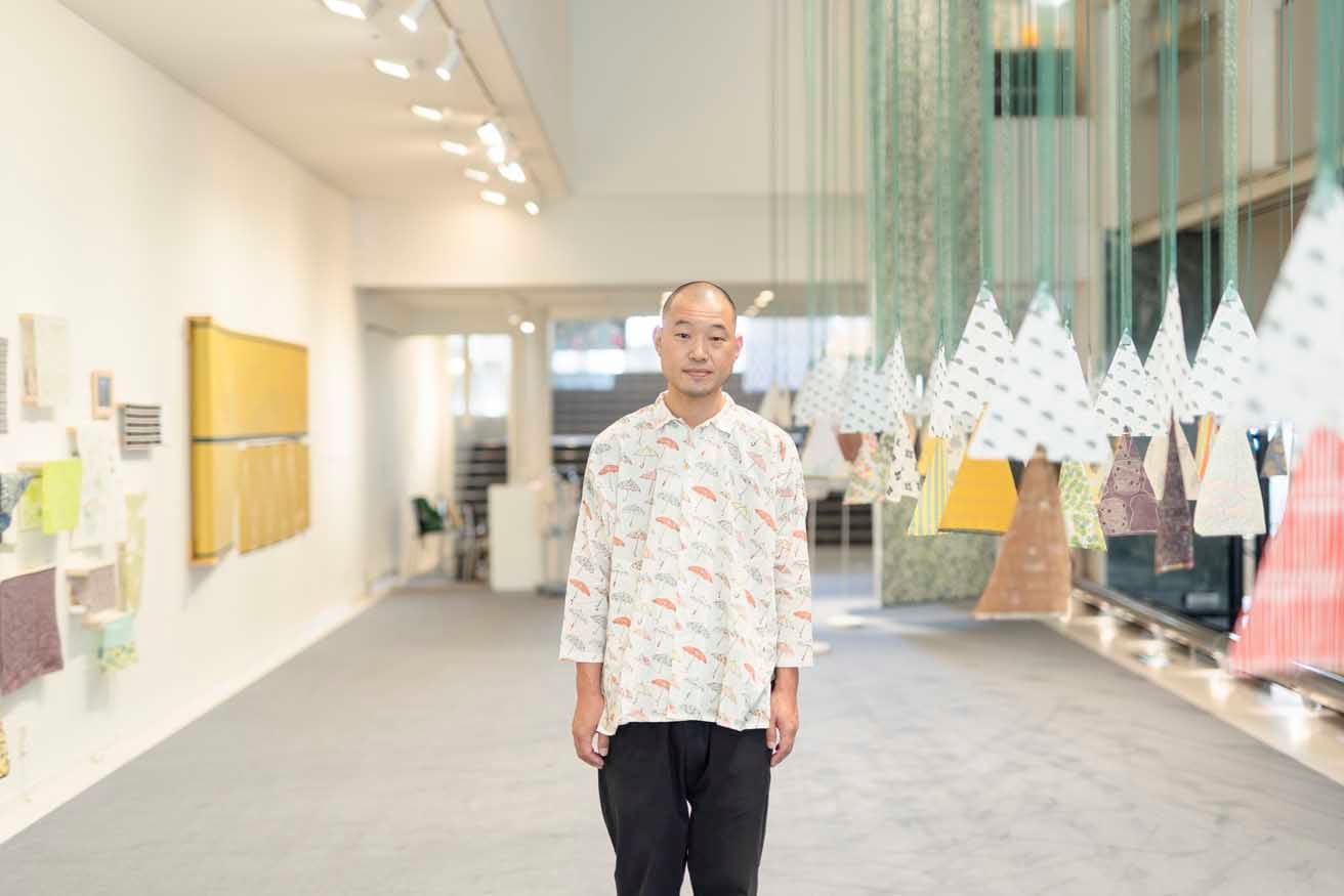 飯田が着用の傘が描かれたシャツはポップアップショップで購入可能。右は100種類のオリジナルテキスタイルに触れて歩きながら、飯田自身によるテーマと技法の解説が読めるインスタレーション「布の森」。photo_Akihiro Yamaguchi
