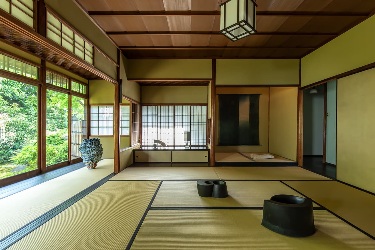 畳敷きの縁側越しに庭園を楽しむ居間。絞り丸太を用いた床と付書院は表千家不審菴の〈残月亭〉を思わせる。地袋上は内田晴之《静止する形》（左）、《Oblong Block Triangular Prism》（右）、縁側は星野曉《Spring Snow 22-S3》、床の間は福本潮子《緑の向こうーII》、植松永次《浮くように》、居間の畳上は手前から秋山陽《抱卵のかたち―壁体》、《抱卵のかたちー双子座》。