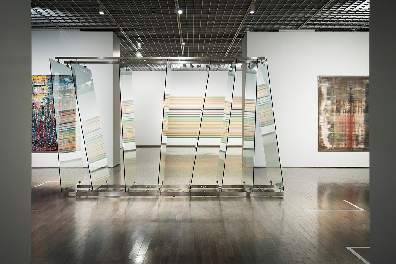 《8枚のガラス》（8枚のアンテリオ・ガラス、スチール／2012／ワコウ・ワークス・オブ・アート蔵）の向こうに隣の展示室の《ストリップ》が見える。